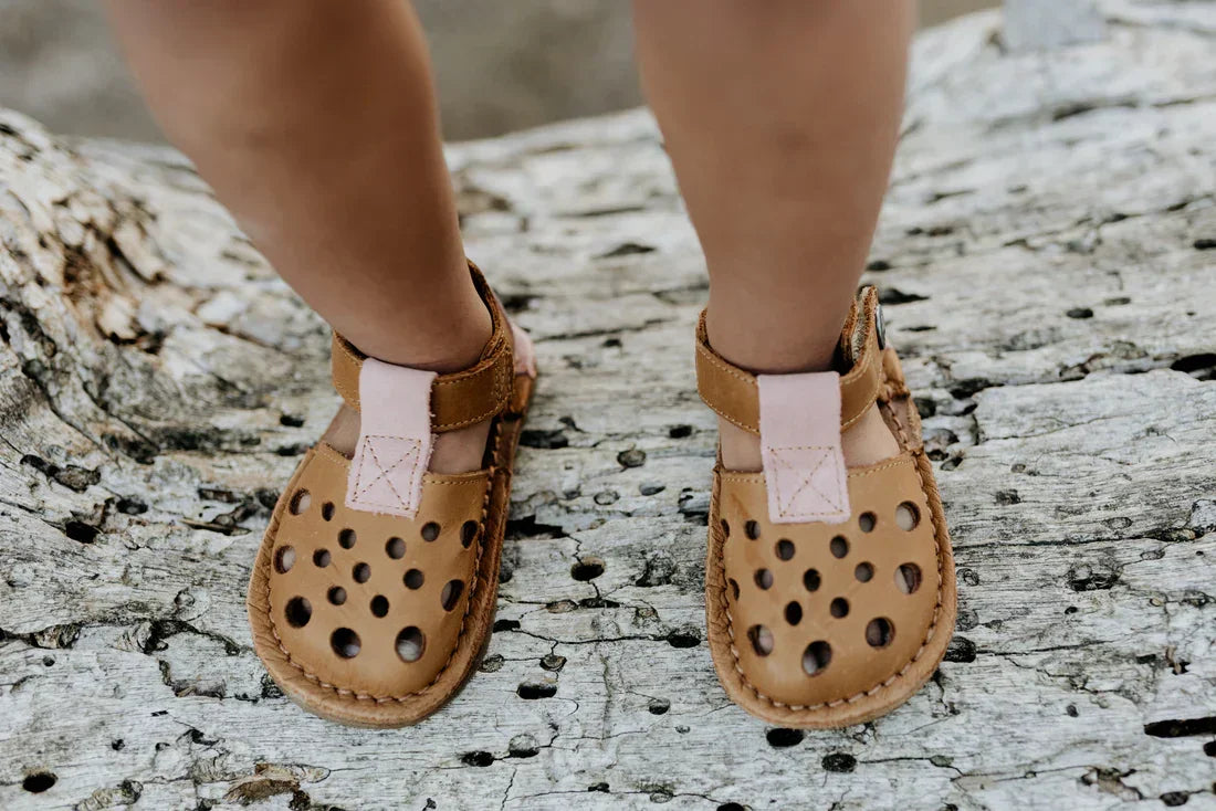 Child wearing amber desert sandals with pink straps standing on weathered wood