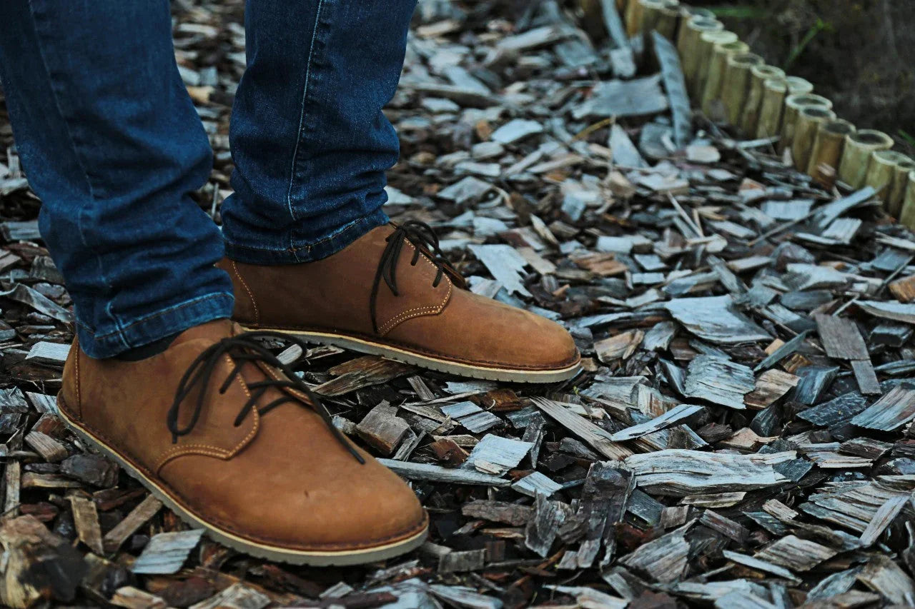Brown natural barefoot shoes on person wearing jeans, standing on wood chips outdoors