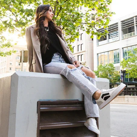 Woman wearing graphite blazer, black top, ripped jeans, and white sneakers sitting on city bench under green trees