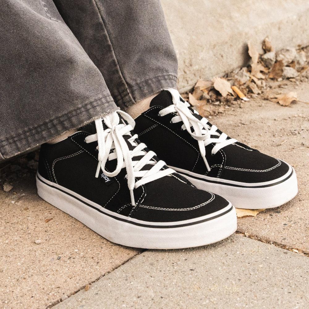 Person wearing black canvas sneakers with white laces and thick white soles on concrete ground with autumn leaves