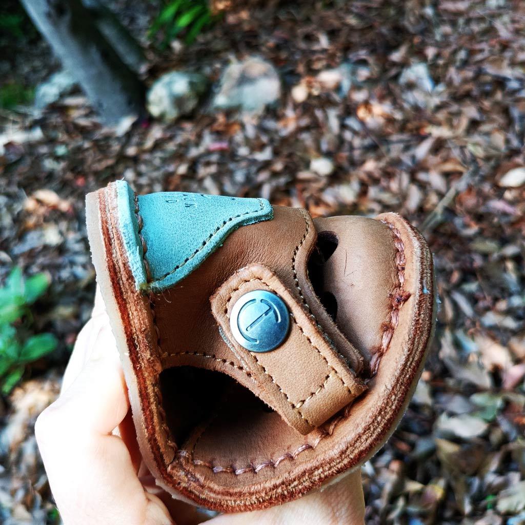 Hand holding a folded brown leather sandal with blue accents and metal button outdoors