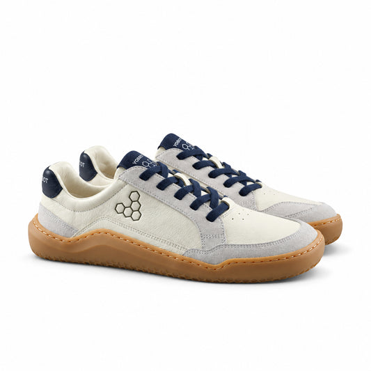 A pair of low-top sneakers in off-white leather with navy blue laces, navy blue heel tab, and a tan gum sole, shown on a light gray background.