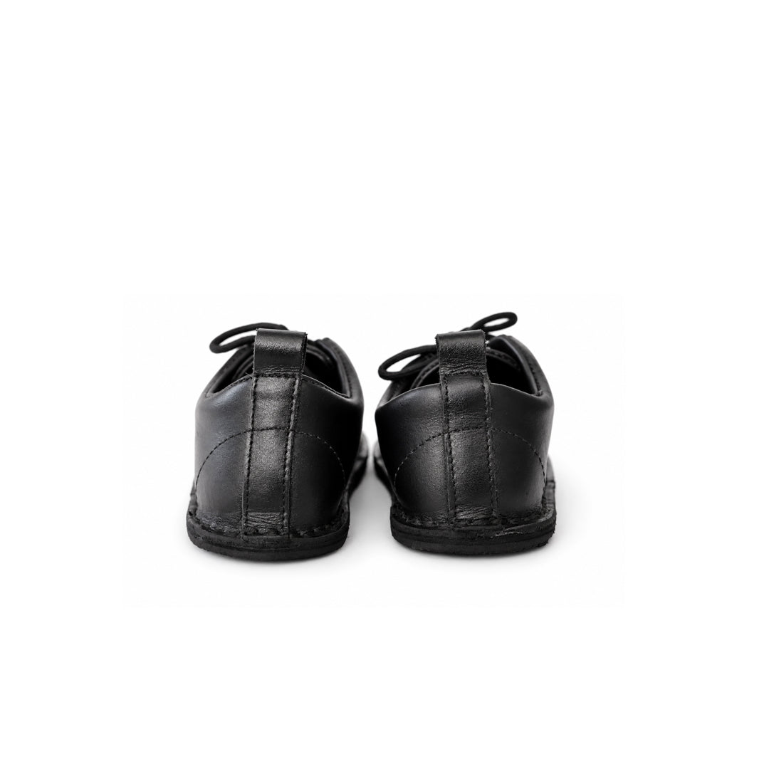 A pair of black leather baby shoes shown from the back, featuring stitched paneling and flat soles, set against a plain white background.