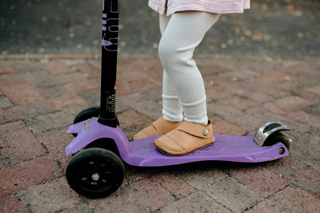 Child wearing beige moccasin shoes standing on a purple three-wheel scooter on a brick pavement