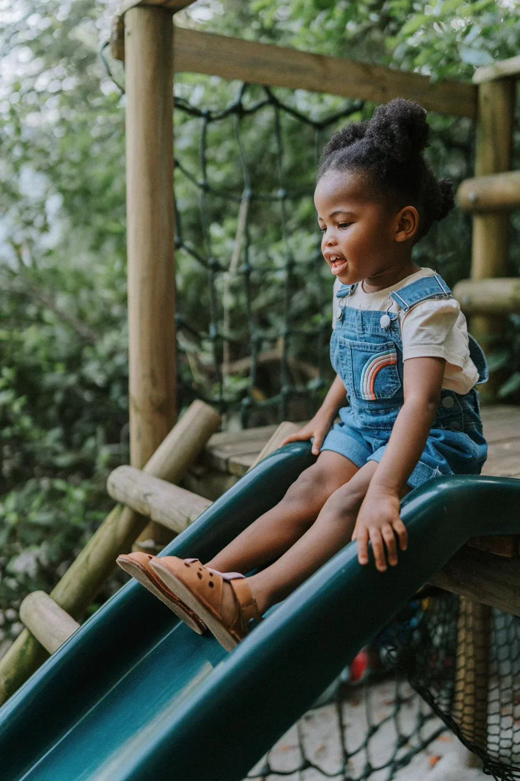 Young girl in denim overalls sliding down a green playground slide outdoors