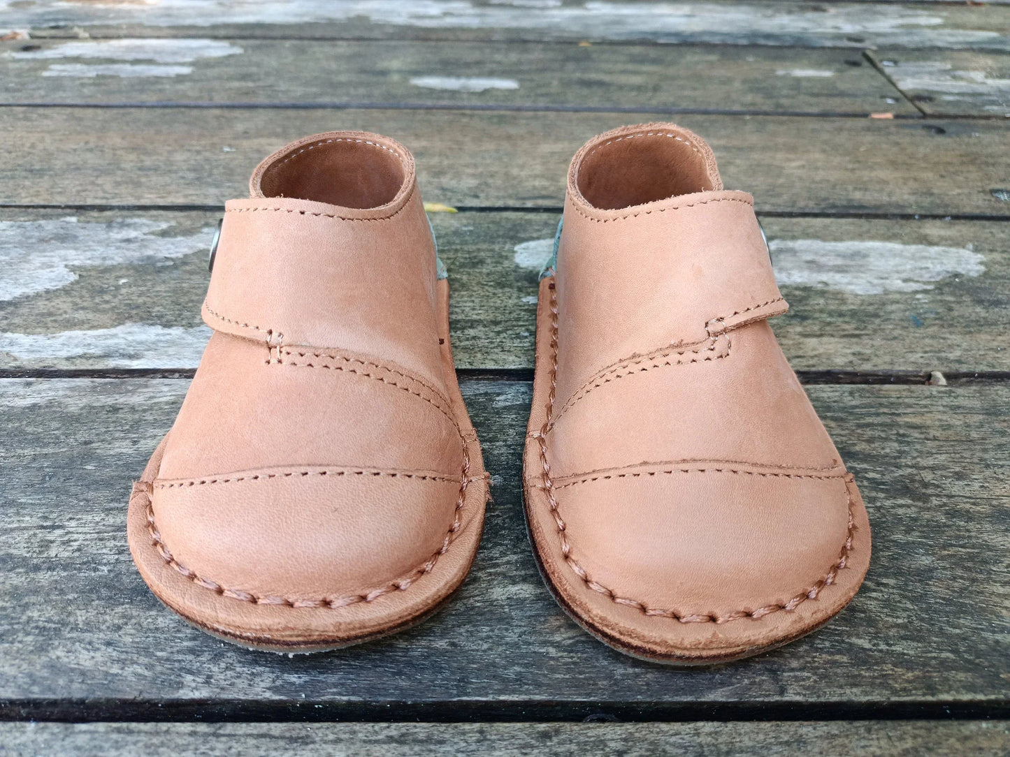 Pair of handmade tan leather baby shoes with stitching detail on rustic wooden floor
