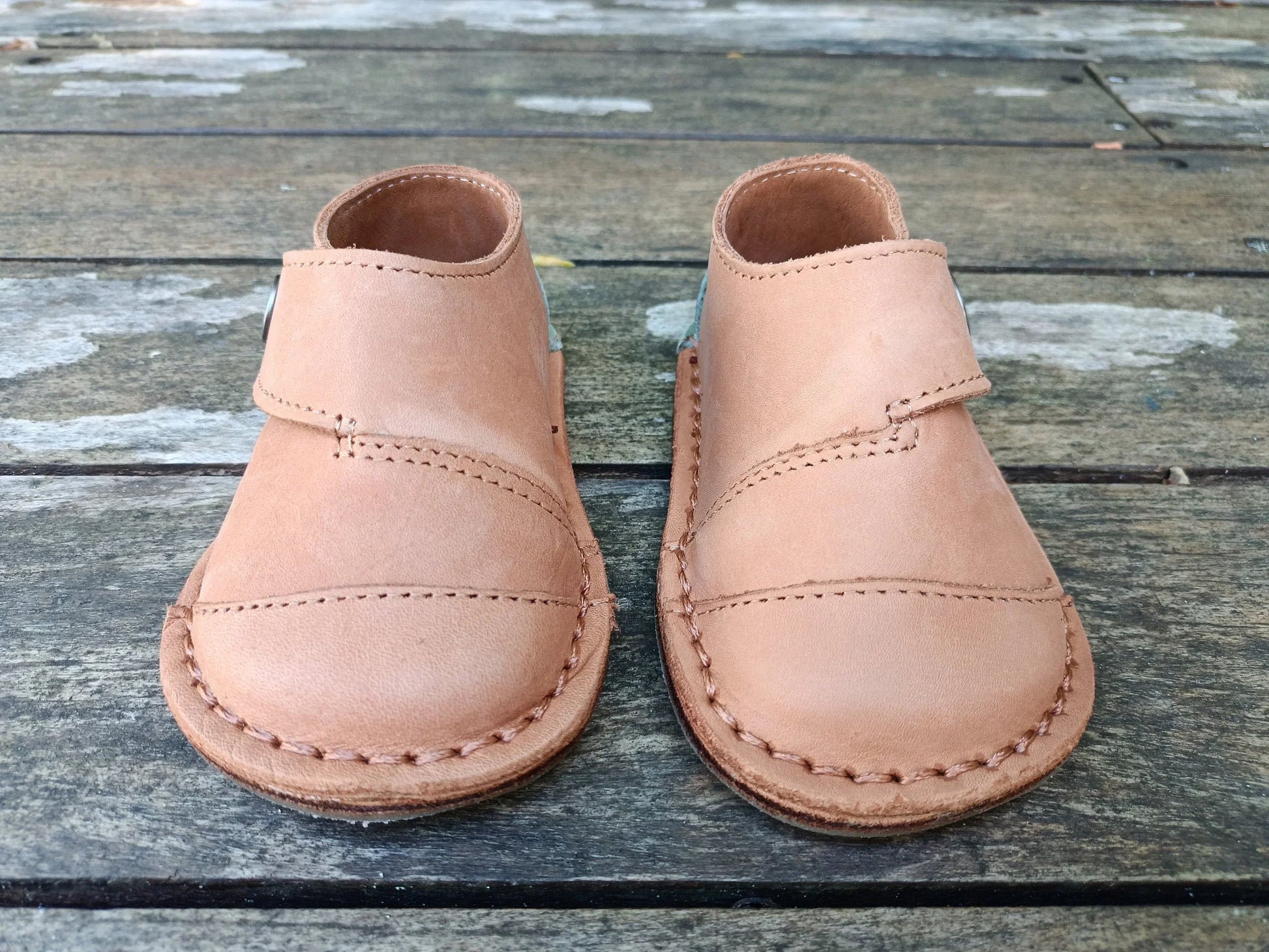 Pair of handmade tan leather baby shoes with stitching detail on rustic wooden floor