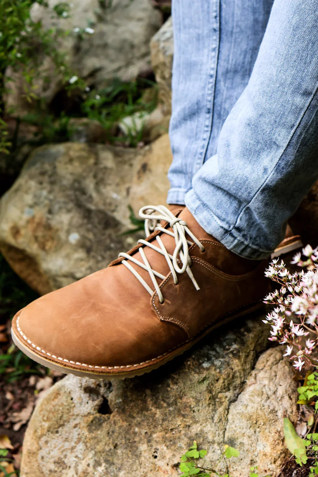 Brown leather casual shoes with white laces worn with light blue jeans on rocky outdoor ground