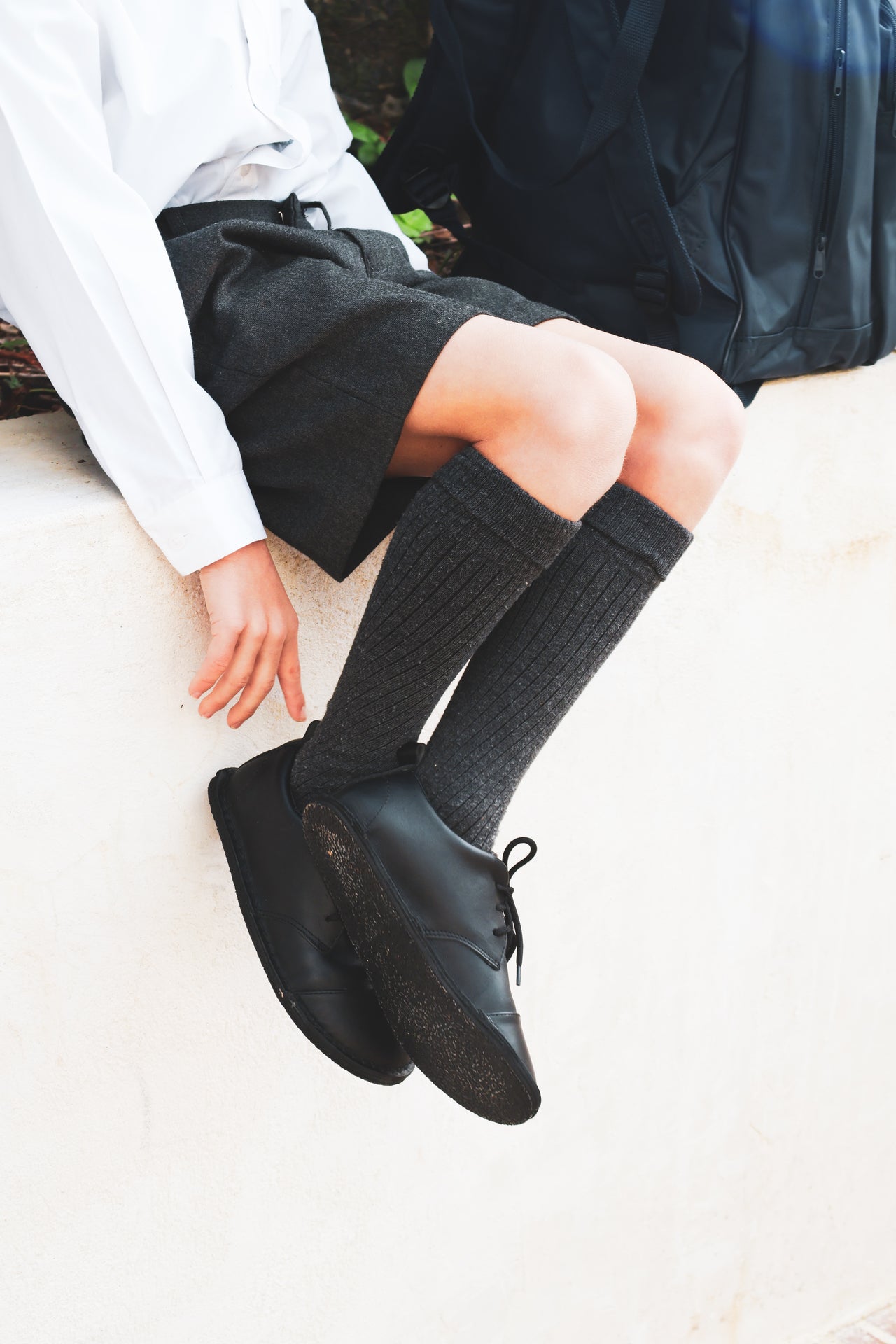 A person sitting outdoors wearing dark gray ribbed knee-high socks, black leather lace-up shoes, a dark skirt, and a white shirt.