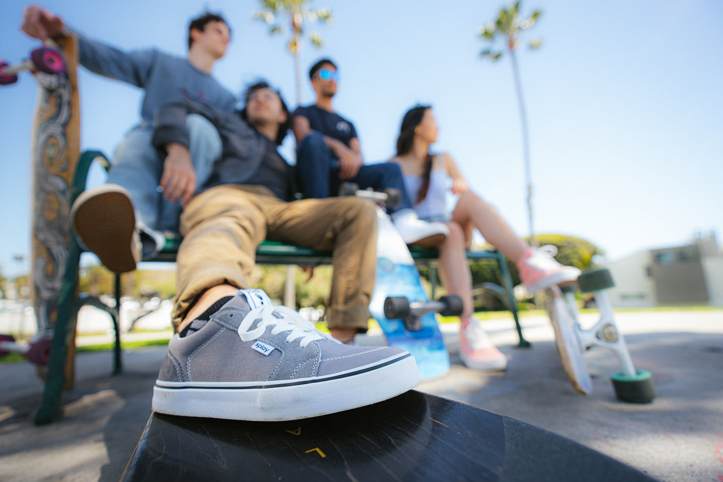 Person wearing a gray shoe with a brand logo, sitting on a skateboard at a skate park with friends in the background.