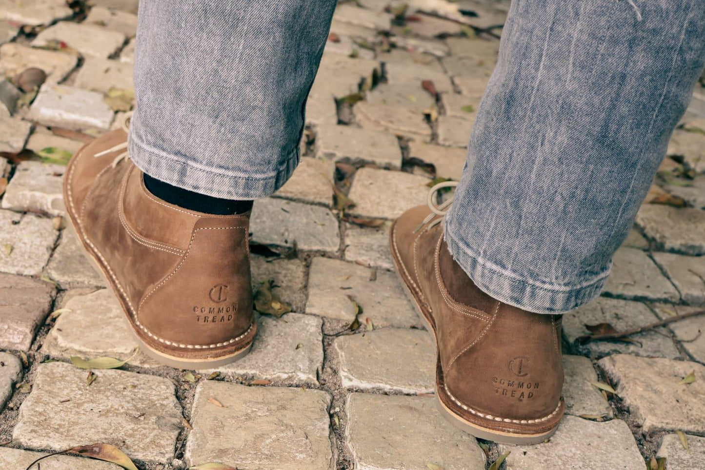 brown Common Tread high cut leather shoes worn with light blue jeans on cobblestone pavement